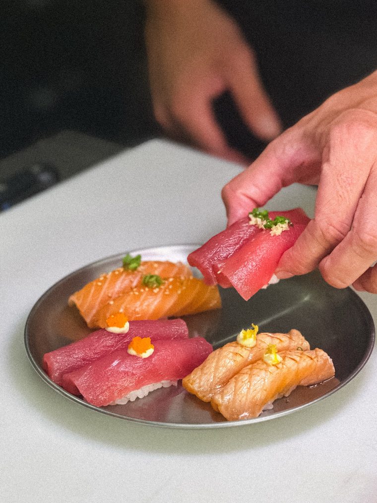 Mão colocando um nigiri de atum num prato de metal já com nigiris de salmão e atum finalizados.