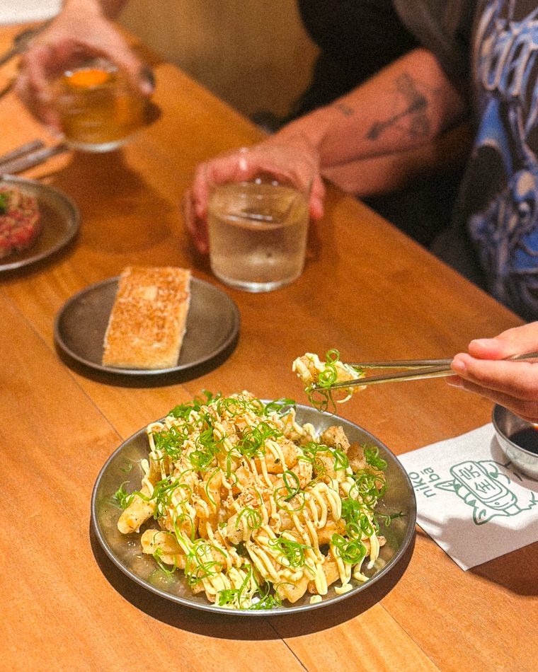 Mesa de madeira no nikō bā com uma travessa de tempurá fries coberta de cebolinha, copos, shokupan tostado e um guardanapo com o gato do nikō impresso.
