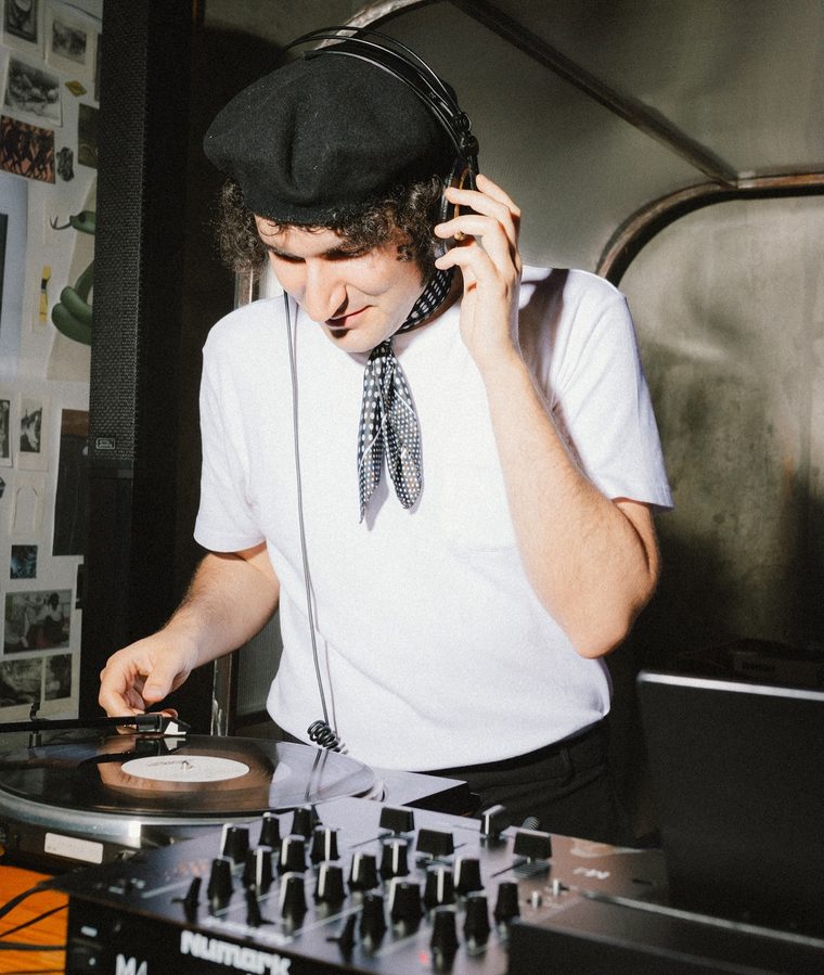 DJ de boina e gravata ajustando o fone atrás da controladora durante o Bailinho.