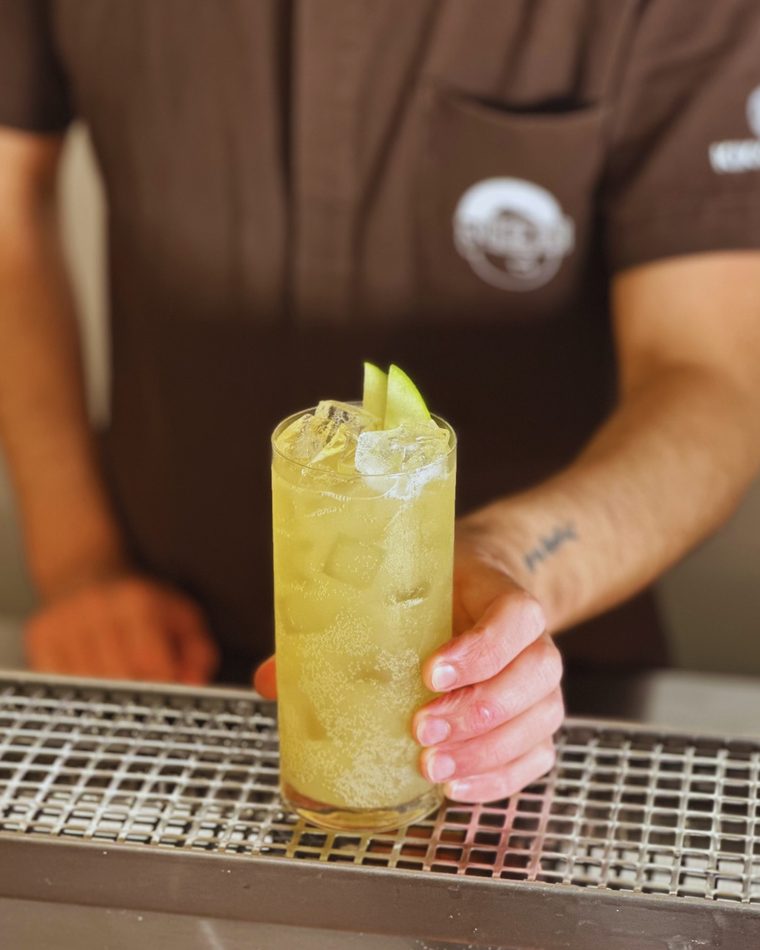 Bartender finalizando um drink alto e gelado com uma fatia de pepino, atrás do balcão do nikō bā.