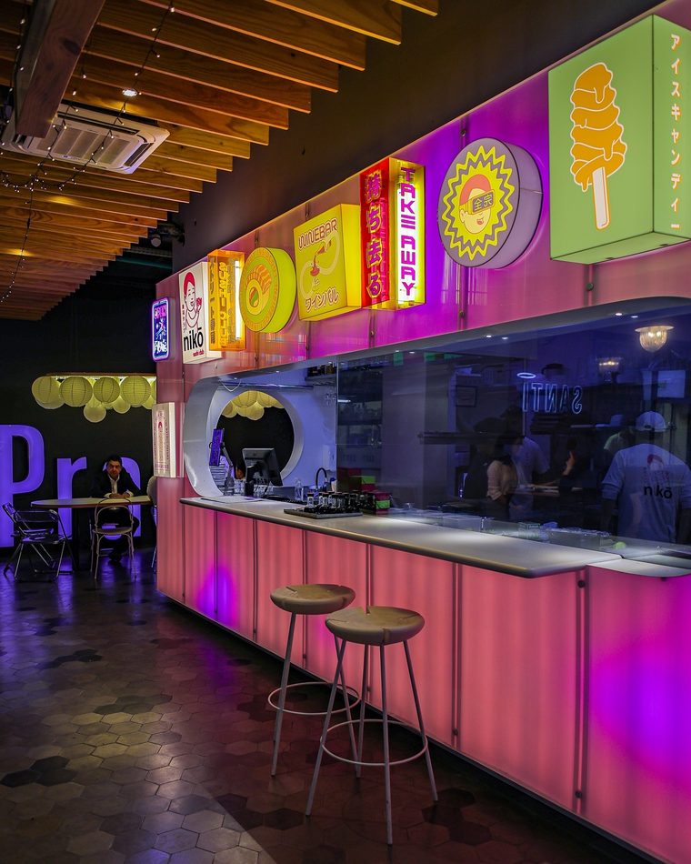 O balcão do nikō sushi club, iluminado em rosa, com os letreiros do cardápio acesos acima da cozinha.