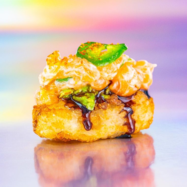 Crispy rice de salmão sobre fundo holográfico: bolinho de arroz frito coberto de tataki de salmão, avocado e molho tarê.