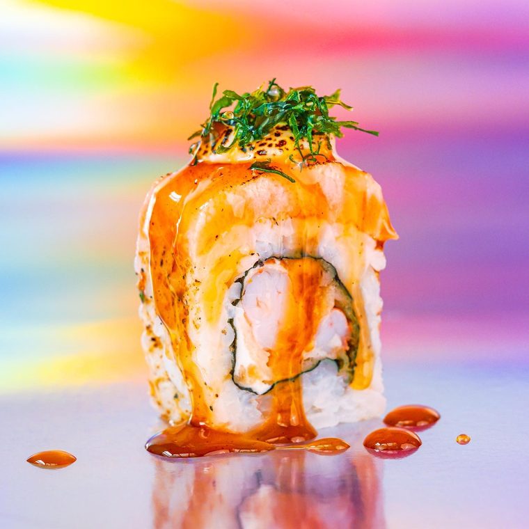 Uramaki Ebi Shake sobre fundo holográfico, coberto por lâmina de salmão maçaricada e molho de laranja.