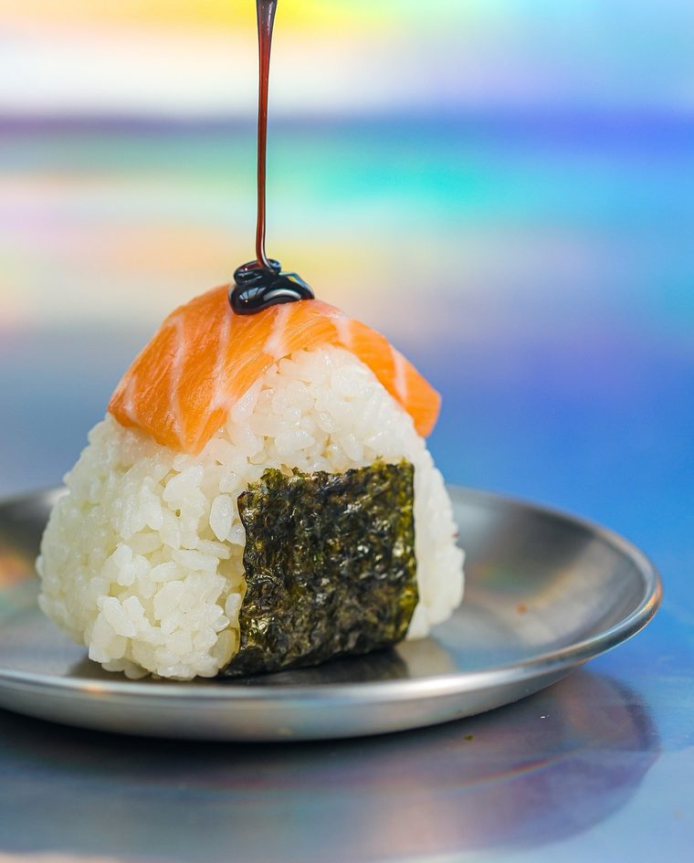 Onigiri Alaska num pratinho de metal, com lâmina de salmão e um fio de molho tarê caindo por cima.