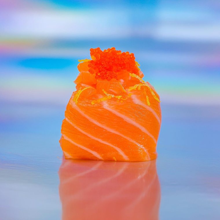 Salmon Joy sobre fundo holográfico: bolinho de arroz envolto em salmão, coberto de ovas de tobiko alaranjadas.