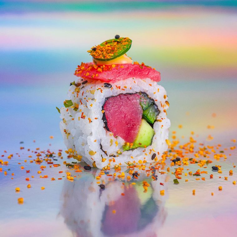 Uramaki Spicy Tuna sobre fundo holográfico, com lâmina de atum, jalapeño e togarashi espalhado ao redor.