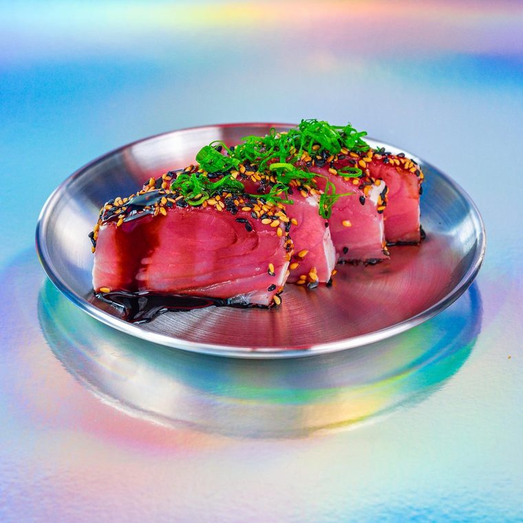 Sashimi de atum na crosta de gergelim, servido num prato de metal iridescente com molho tarê e cebolinha.